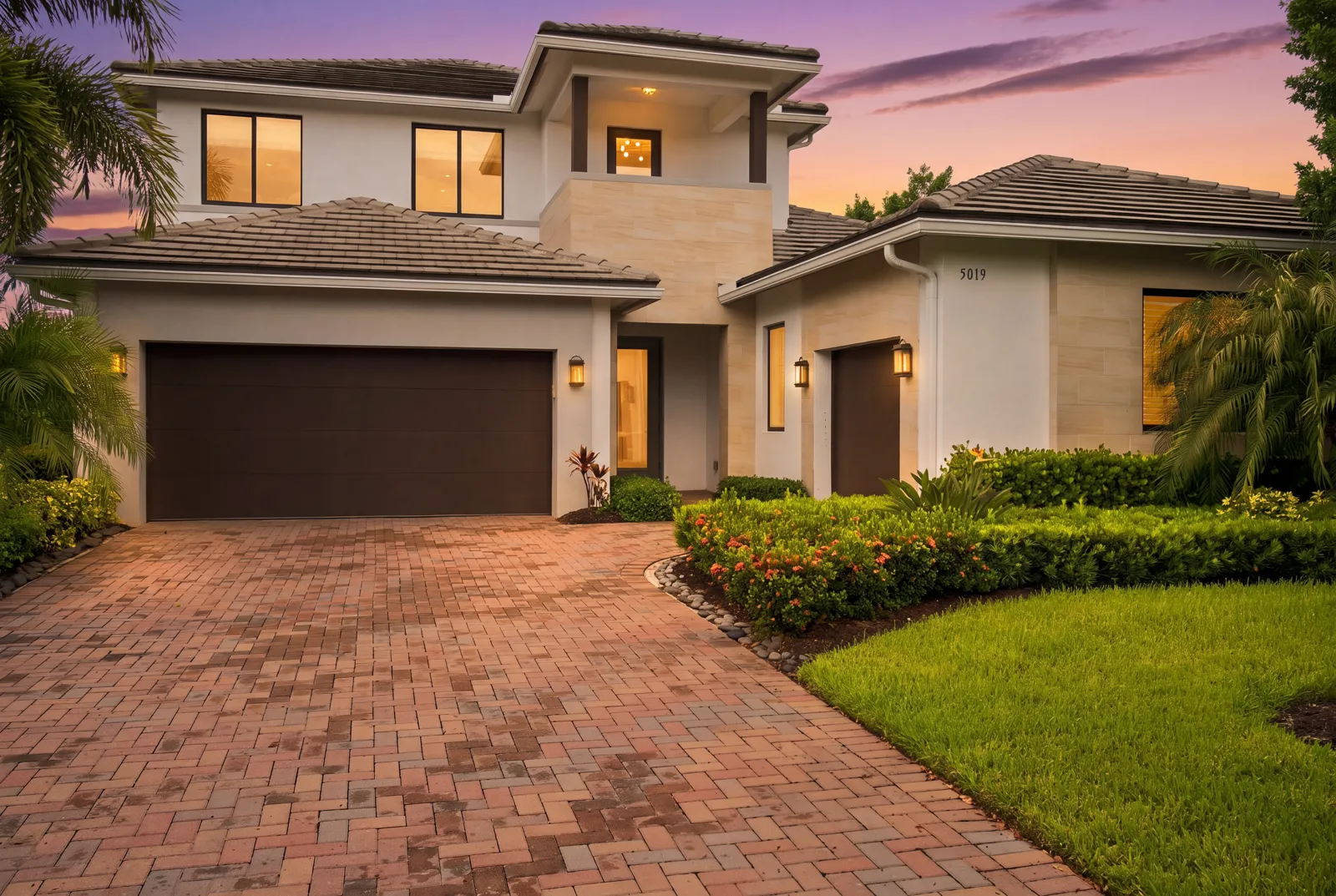 Two-story home at dusk with warm lights along a brick paver courtyard (virtual twilight, digitally enhanced)